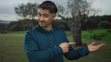 Serin bir park ortamında avucunun içini gösterip el hareketi yapan genç İspanyol bir adam..