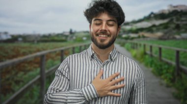 Sakallı genç adam yeşil bir parkta duruyor. Gülümsüyor ve göğsüne dokunuyor. Bulutlu bir gökyüzünün altında huzurlu doğal ortamın tadını çıkarıyor..