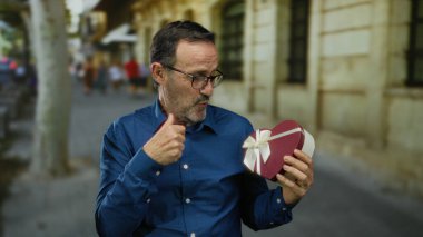 Elinde kalp kutusu olan yaşlı bir adam açık şehir parkında bulanık taş bina arka planıyla anlamlı bir şekilde işaret ediyor..