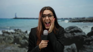 Sahilde muhabir olarak mikrofon tutan bir kadın gözlüklü açık havada okyanus manzarası karşısında şaşırmış görünüyor..