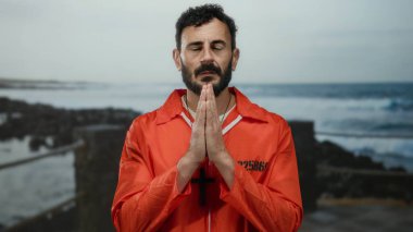 Turuncu tulumlu bir adam kumsalda dua ediyor. Arka planda okyanus dalgaları var. Bulutlu bir gökyüzünün altında siyah bir haç kolye takıyor ve dikkatlice yukarı bakıyor..