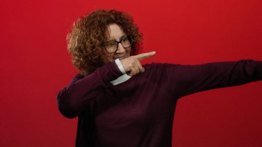 Kıvırcık saçlı ve gözlüklü, kendine güveni tam bir jest sergileyen canlı kırmızı arka planda gülümseyen bir kadın..