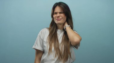 Beyaz tişörtlü İspanyol kadın, izole edilmiş mavi arka plan duvarında boyun ağrısını dile getiriyor ve gerçek bir rahatsızlık anı yakalıyor..