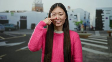 Şehir caddesinde duran pembe kazaklı bir kadın burnuna gülümseyen bulanık şehir arka planıyla açık hava manzarasını tasvir eden..