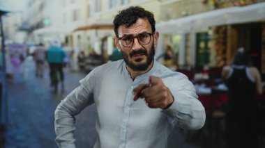 Canlı sokak sahnesinde sakallı İspanyol bir adam masaların ve yayaların hareketli kentsel geçmişleri arasında gözlük ve gri gömlek takıyor..