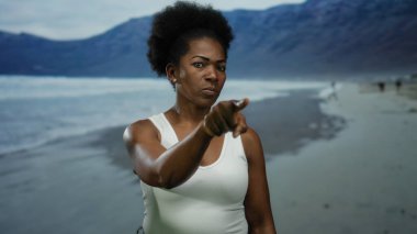 Sahilde el kol hareketleri yapan, arka planda dağlar olan bir kadın açık havaya davet ediyor..