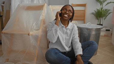 Oturma odasında telefonla gülümseyen bir kadın. Yeni ev eşyalarıyla çevrili. Mutluluk ve modern yaşam tarzını gösteriyor..