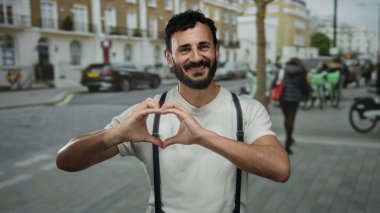 Sakallı İspanyol bir adam gülümsüyor ve elleriyle şehir dışındaki bir sokakta bisiklet ve arka plandaki insanlarla kalp oluşturuyor..