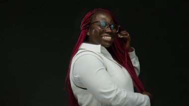 Gözlüklü, örgülü saçlı bir kadın izole edilmiş siyah bir arka plana gülümsüyor neşeli bir ifade ve profesyonel giysiler sergiliyor.