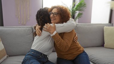 Kadın arkadaşlar rahat bir oturma odasındaki kanepede kucaklaşıyorlar, rahat bir iç mekanda sıcak bir anı paylaşıyorlar, yakın ilişkilerini ve neşeli arkadaşlıklarını yansıtıyorlar..