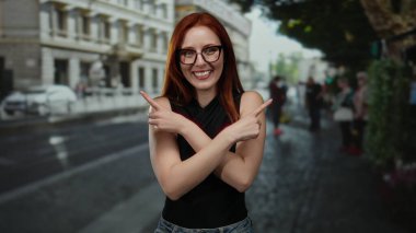 Bir şehir caddesinde kollarını açmış gülümseyen bir kadın, kırmızı saçlarını ve gözlüklerini şehir zemininde sergiliyor, canlı bir açık hava atmosferi yakalıyor..