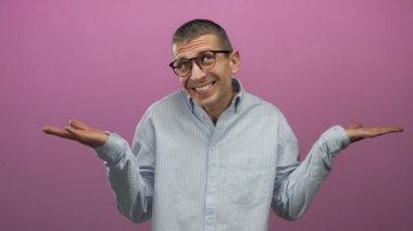 Gözlüklü, çizgili tişört giyen beyaz bir adam pembe arka plana karşı duruyor. Omuz silkme, izole ve gayri resmi bir ortam ile kafa karışıklığı gösteriyor..