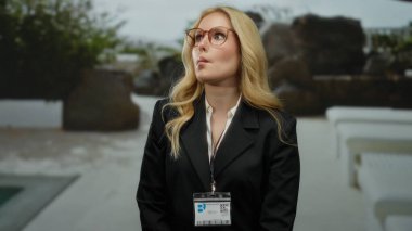 Otelin yanında yüzen, gözlüklü, siyah takım elbiseli, açık hava ortamında sarı saçlı bir kadın eğlenceli bir ortam yaratıyor..