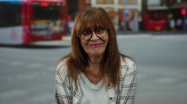 Arka planda bulanık otobüsler ve ekose gömlek ve gözlüklerle işlek bir şehir caddesinde neşeli bir şekilde gülümseyen İspanyol son sınıf öğrencisi kadın..