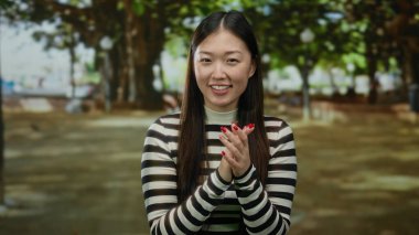 Açık havada gülümseyen bir kadın, güneşli bir parkta yeşil ağaçlı çizgili bir süveter giyiyor, neşeli bir ifade ve doğal güzellik sergiliyor canlı, huzurlu bir açık hava ortamında..