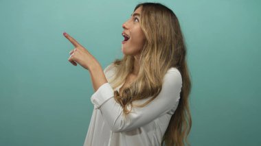 Genç sarışın kadın stüdyo ortamında yukarı doğru işaret ediyor, açık ağızlı ve geniş gözlü çerçeveden dışarı bakıyor; sürpriz merak şaşkınlık.