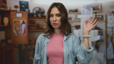 İçerideki Latin kökenli genç kadın, elini kaldırmış, rahat bir odada, arka planda rafları olan tarafsız bir yüz ifadesiyle güvenle dikiliyor..