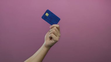 Kafkasyalı bir adamın elinde pembe bir duvara karşı mavi bir kredi kartı tutarken, canlı bir arka planda izole edilmiş, finans ve kişisel kredi kullanımını vurgulayan.