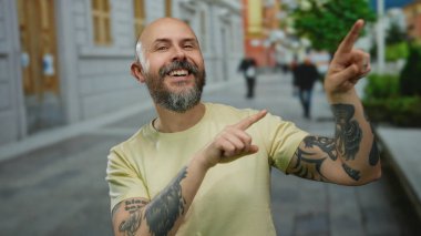 Şehirde gündüz vakti sakallı ve dövmeli bir adam açık havada kel kafasıyla mutlu bir yüz ifadesi sergiliyor..