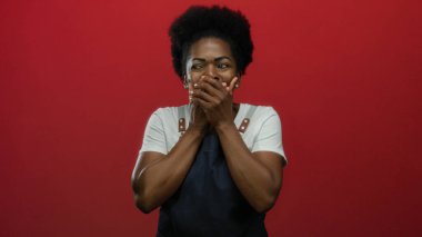 Kırmızı arka planda önlüklü genç bir kadın, ellerini şaşkınlık ve neşe içinde dudaklarını kapatıyor, duygusal ve ilgi çekici bir an sergiliyor..