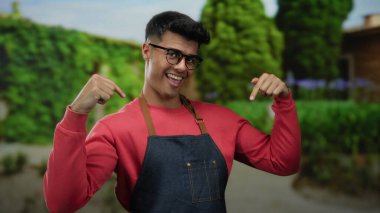 Gözlüklü ve önlüklü genç adam gülümsüyor ve yeşillik ve patikalarla çevrili bir açık hava parkına işaret ediyor..