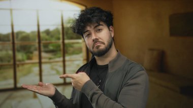 Salondaki sakallı genç adam avucunun içini işaret ederken geniş pencereden doğa görüşü ile düşünceli bir görünüm sergiliyor ve iç gözlemci bir atmosfer yaratıyor..