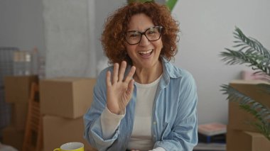 Oturma odasında gülümseyen bir kadın. Etrafı kutularla çevrili. Yeni ev heyecanını yansıtıyor..