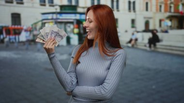 Şehir caddesinde İsveçli banknotlarla gülümseyen bir kadın, arka planda tarihi binalar bulunan açık şehir ortamında kızıl saçlı güzelliği sergiliyor..