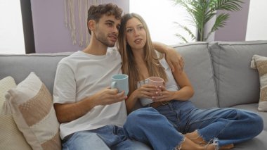 Sıcak oturma odasındaki kanepede dinlenen bir çift, ev ortamında sıcak içeceklerin tadını çıkaran bir erkek ve bir kadın, şık iç dekorun ortasında sıcaklığı ve aşkı iletiyorlar..