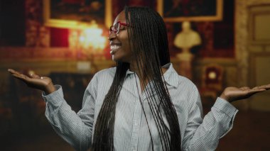 Kapalı müze ortamında kollarını açmış gülümseyen bir kadın, gözlüğünü ve örgülü saçlarını zarif bir arka planda örüyor..