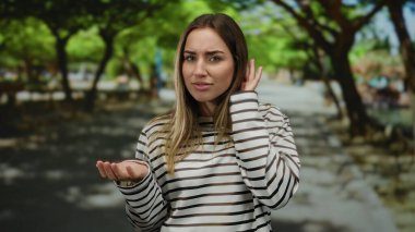 Sarı saçlı bir kadın güneşli bir parkta yeşil ağaçlarla çevrili, çizgili bir gömlek giymiş açık hava manzarasını ve neşeli havasını vurgulayan bir jest yapıyor..