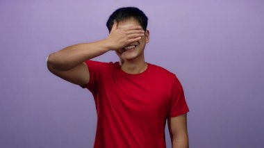 İzole edilmiş mor arka planı olan yüzü kaplayan genç bir adam kırmızı bir gömlek giyiyor, neşeli ve canlı bir ifade sergiliyor..