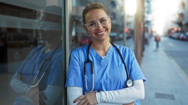 Steteskoplu gülümseyen kadın hemşire gündüz vakti şehir kliniğinin önünde güvenle duruyor..