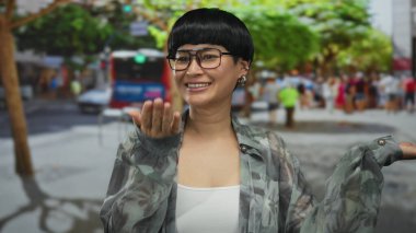 Kentsel sokak ortamında gülümseyen, el kol hareketi yapan şehir ışıkları altında neşeli bir ifade sergileyen, arabaların bulanık arka planı ve canlı metropoldeki yeşilliklerle çerçevelenmiş bir kadın..