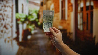 Elinde tekil bir banknot tutan, şehir arkaplanını gözler önüne seren, şehir dışında yatırım temalarını ön plana çıkaran bir adamın eli..