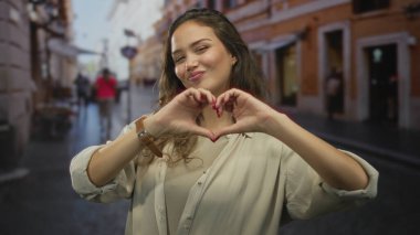 Açık havada gülümseyen bir kadın, şehir caddesinde kalp işareti yaptırıyor, şehirde mutlu bir ifadeyle giyinmiş, sıcaklığı ve arkadaşlığı iletiyor..