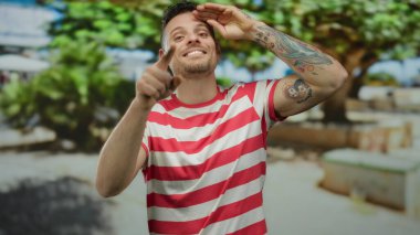 Genç adam gülümsüyor ve güneşli bir günde çizgili bir gömlek giyerken şehir ortamında başparmağıyla işaret ediyor.