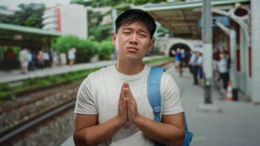 Tren istasyonunda mavi sırt çantalı genç bir adam, kalabalık bir açık hava ortamının ortasında yalvaran bir jest yapıyor..