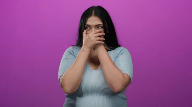 Kadın elleri pembe arka planda durarak ağzını kapatıyor, şaşkınlığını ifade ediyor, mavi gömlek giyiyor, duygularını ifade ediyor, genç, iri yarı bir kadına ait, canlı bir ortamda izole edilmiş..