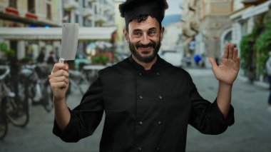 Sakallı, siyah üniformalı İspanyol aşçı balta tutuyor açık havada, hareketli bir sokakta gülümsüyor..