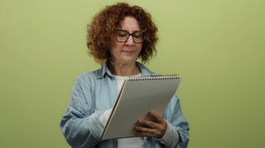 Middle-aged woman writing in notebook against isolated yellow background, wearing glasses and casual denim shirt.