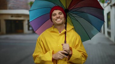 Sarı yağmurluklu adam şehir caddesinde renkli bir şemsiye tutuyor.