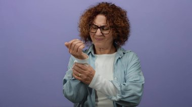 Mor duvarın önünde bileği ağrıyan bir kadın gözlük takıyor ve mavi bir gömlek giyiyor. Yüzünde endişeli bir ifade var. İçeriden izole edilmiş..