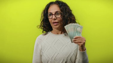 Orta yaşlı bir kadın elinde parlak sarı bir arkaplan karşısında sürpriz bir ifadeyle uae dirhams tutuyor.