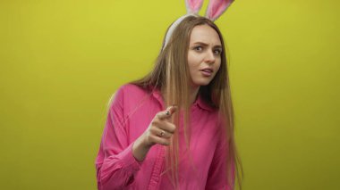 Tavşan kulaklı bir kadın, pembe gömlekli, sarı bir arka plana neşeli bir şekilde işaret ediyor. Genç, kaygısız bir havayla eğlenceli bir paskalya teması öneriyor..