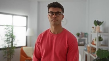 Gözlüklü ve kırmızı kazaklı genç adam, modern iç mekan yaşam tarzını ön plana çıkaran bitkiler ve bebek dekoruyla süslenmiş rahat bir yatak odasında düşünceli bir şekilde duruyor..