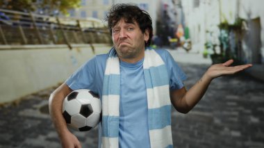 İspanyol asıllı bir adam sokakta futbol topunu tutuyor. Sıradan kıyafetler ve mavi eşarpla şaşkın şaşkın bakıyor..