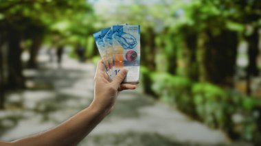 Şehir caddesinde İsviçre frangı banknotları tutan bir adam, şehir merkezini ve finansal temayı tasvir ediyor, bulanık yaya geçmişiyle şehir hayatını vurguluyor..