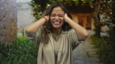 Genç İspanyol kadın dışarıda elleri kulaklarının üzerinde hayal kırıklığını ifade ediyor. Manzaralı bir bahçe yolunda duruyor. Bej bir kıyafetle stres ve gerilim duygularını gösteriyor..