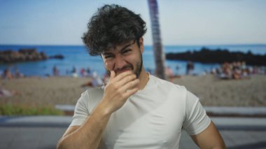 Sahilde sakallı genç İspanyol bir adam hapşırıyor, arka planda palmiye ağacı ve deniz görünüyor, açık deniz sahili anını yakalıyor..